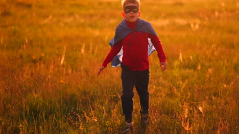 A-boy-in-a-suit-and-a-superhero-mask-with-a-red-cloak-runs-across-the-field-at-sunset-on-the-grass-dreaming-and-imagining-himself-a-hero.